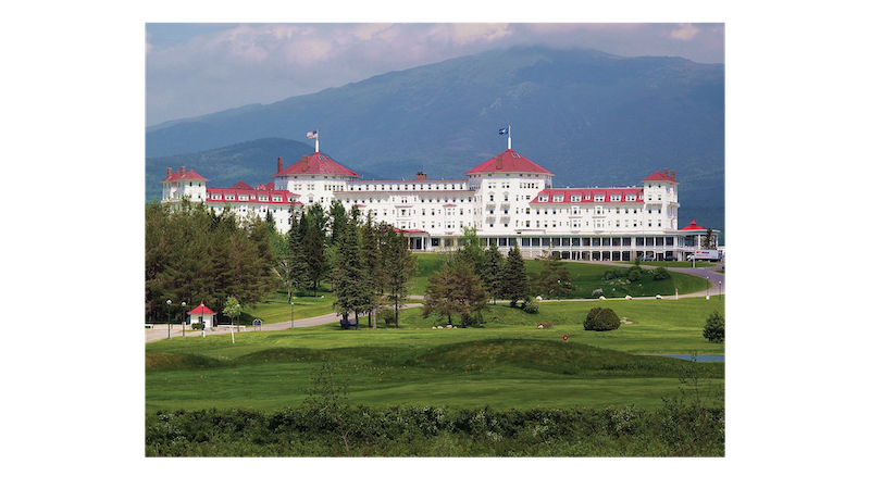 a large building foregrounded by rolling green hills and trees, backgrounded by a small mountain